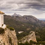 The belltower of Guadalest perched on top of a cliff with the valley running down to the sea, and dark storm clouds in sky above.